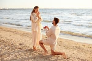 love and people concept - smiling young man with engagement ring making proposal to happy woman on beach