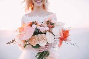 Close-up of a bride’s bouquet of amaryllis, anthurium, roses, carnations, eucalyptus in white-peach shades. The bride in a lace white dress holds a bouquet in her hands. White sand