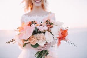 Close-up of a brideâ€™s bouquet of amaryllis, anthurium, roses, carnations, eucalyptus in white-peach shades. The bride in a lace white dress holds a bouquet in her hands. White sand