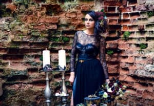 A girl in a mystical dark image in a black dress with flowers in her hair against the background of an old brick wall. Candles, black magic, Gothic beauty, mystical image. Halloween, black widow