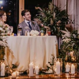 Luna + Lion Photo Co. Couple sitting at sweetheart table surrounded by leafy green arrangements and Pampas grass